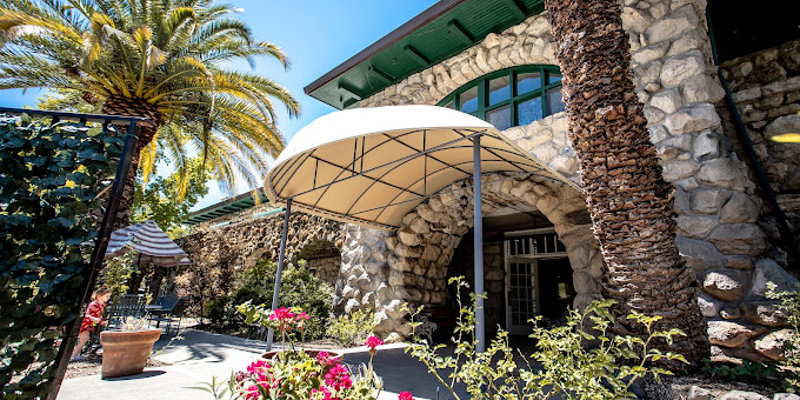  A charming stone building framed by tall palm trees and vibrant flowers, featuring a welcoming canopy entrance under a bright blue sky.