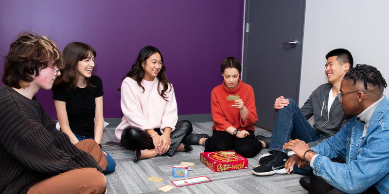 A group of young adults sits together on the floor, laughing and enjoying a board game in a relaxed, friendly setting with a purple accent wall.