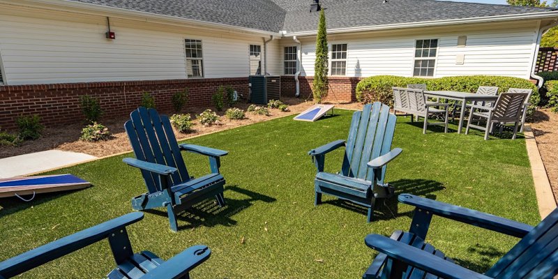 A cozy outdoor space with blue Adirondack chairs on a grassy lawn, surrounded by vibrant greenery, perfect for relaxation and games.