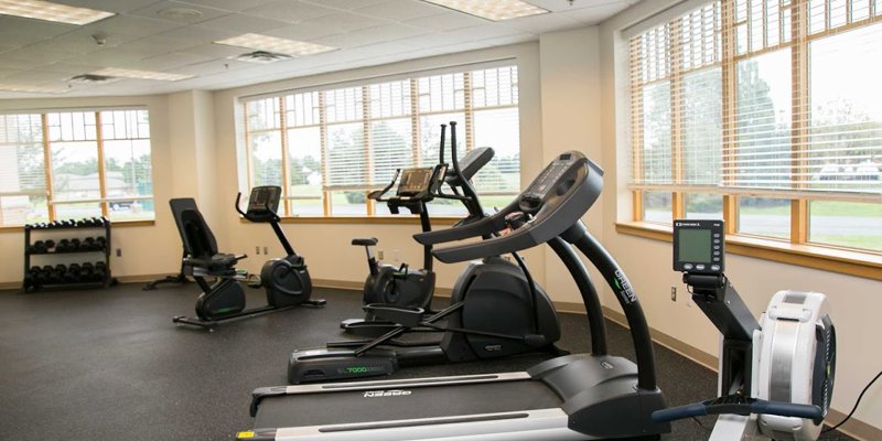 A bright and spacious fitness room equipped with treadmills, exercise bikes, weights, and large windows offering natural light and outdoor views.