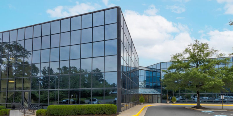  A sleek, modern glass building with reflective windows, surrounded by greenery and a well-maintained parking area under a bright, clear sky.