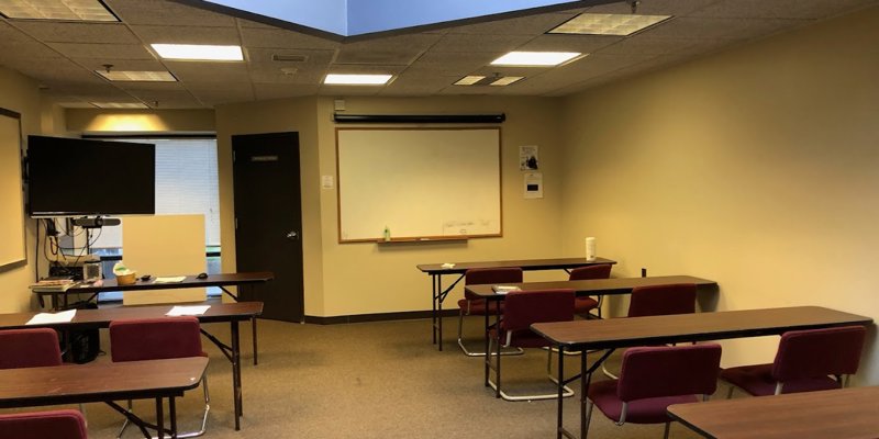 A cozy, small classroom with burgundy chairs and tables arranged for a meeting or lecture. It features a whiteboard, a TV, and a skylight.