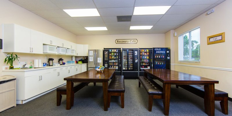 A welcoming dining area labeled "Recovery Café" with large wooden tables, benches, vending machines, and a fully equipped kitchen, offering a cozy space for meals.