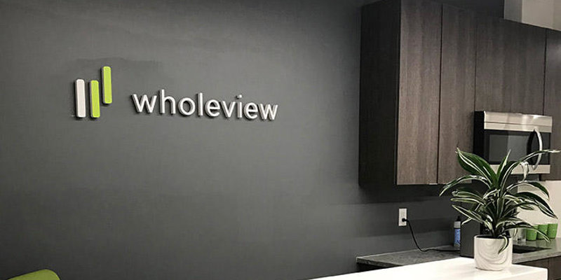 A sleek, modern reception area with a clean design, featuring the "wholeview" logo on a dark wall, a white counter, and a potted plant.