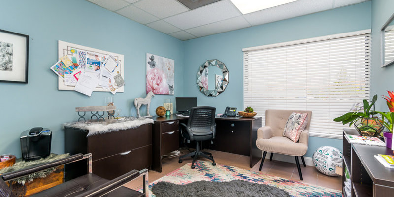 A bright and stylish office with a soft rug, modern desk, cozy seating, and plants. The decor includes a bulletin board and floral artwork for a calming vibe.