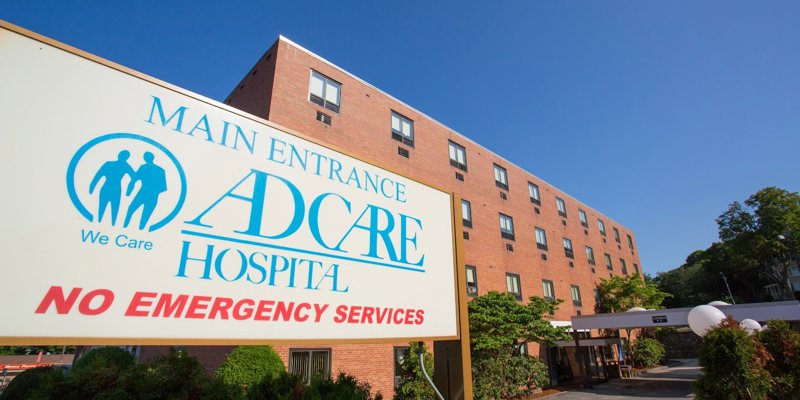 Adcare Hospital's entrance is prominently displayed under a clear blue sky, with a welcoming sign, well-maintained landscaping, and clear directions.