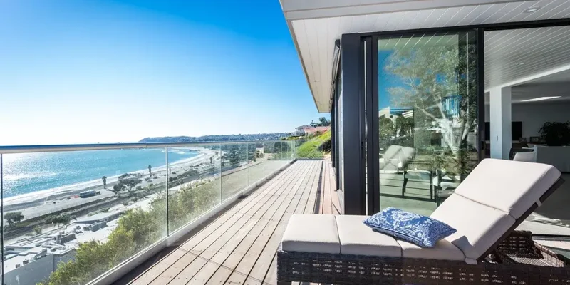 Sunlit coastal retreat with a spacious deck, modern lounge chairs, and glass railing, overlooking a serene beach and blue ocean views.