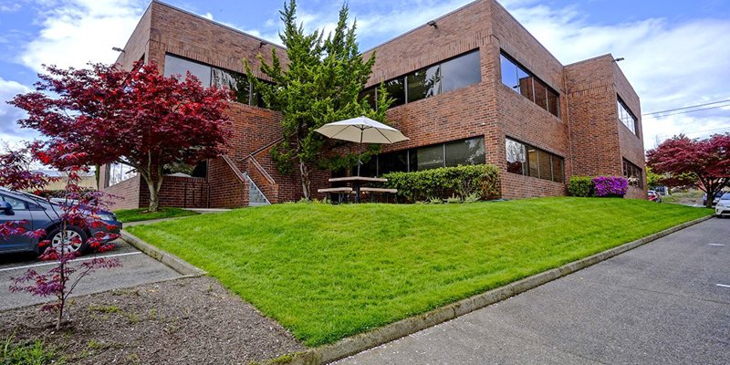 A modern brick building with large windows, surrounded by well-maintained greenery and vibrant trees. A picnic table sits under an umbrella.