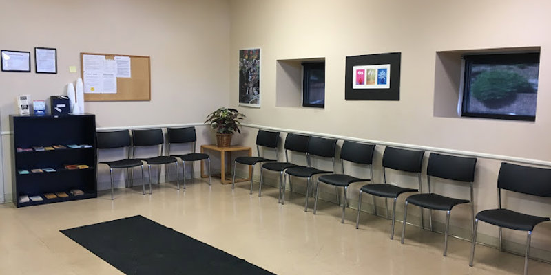 A clean, organized waiting room with rows of black chairs, a bulletin board, and neatly displayed brochures. Natural light enters through small windows.