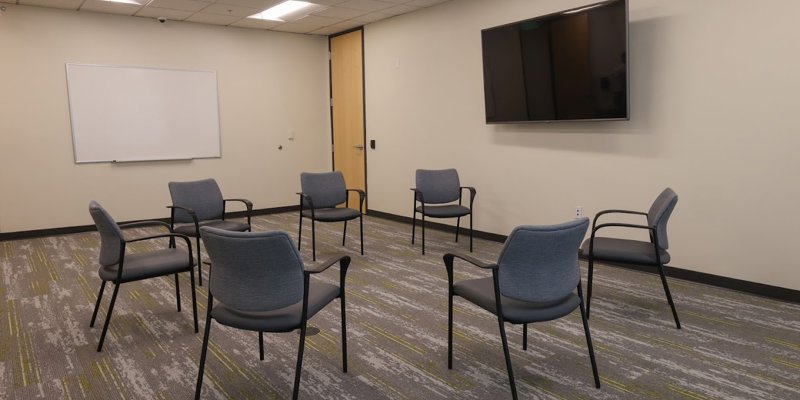 A well-lit group therapy room with a circular arrangement of comfortable chairs, a large TV screen, and a whiteboard for discussions.