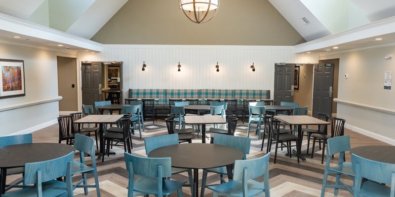 A bright, spacious dining area featuring modern round tables, blue chairs, and a high ceiling with a large, elegant chandelier.