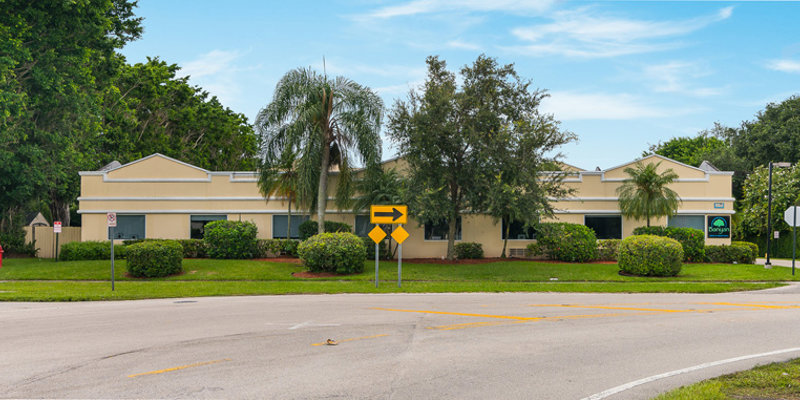 A well-maintained one-story building surrounded by lush greenery and palm trees at a quiet intersection, featuring clear directional signage.