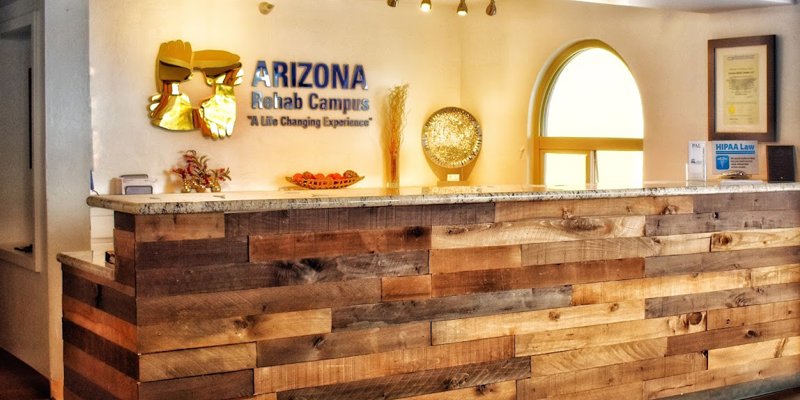 A welcoming reception desk made of rustic wood sits beneath soft lighting, with a sign displaying "Arizona Rehab Campus" and a warm, inviting atmosphere.