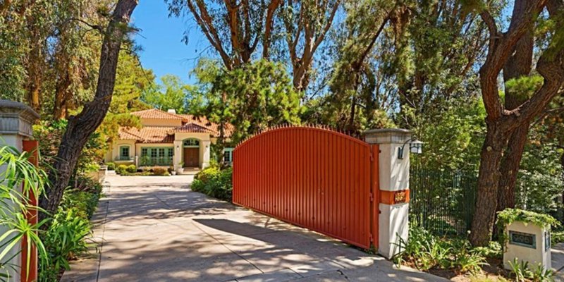  A charming Mediterranean-style home is tucked behind a bright red gate, surrounded by lush trees and greenery, offering privacy and elegance.