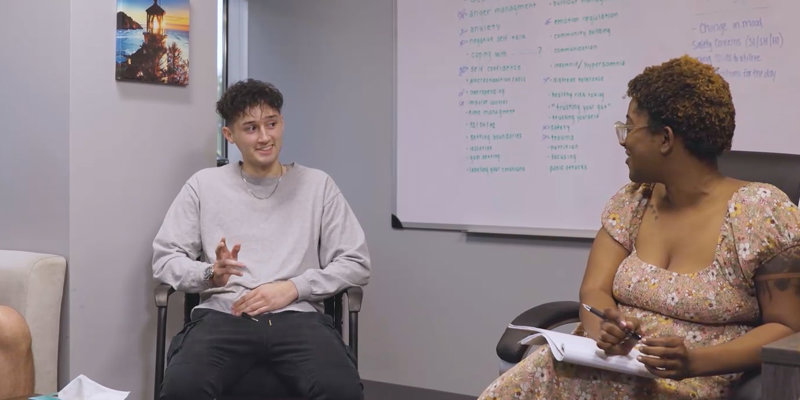 Two individuals are engaged in a conversation in a well-lit room, with a whiteboard featuring various discussion points in the background.