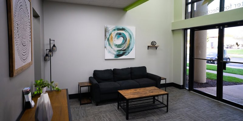 Modern and inviting waiting area with a comfortable black sofa, abstract artwork, wooden furniture, and large windows for natural light.