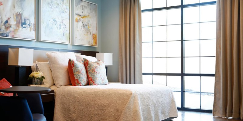 A serene bedroom with elegant decor features a cozy bed adorned with vibrant pillows, modern art, and large windows that flood the space with natural light.