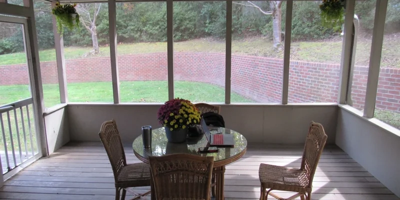 A serene screened-in porch with wicker chairs around a glass table, overlooking a lush, green yard with a brick wall, creating a peaceful outdoor retreat.