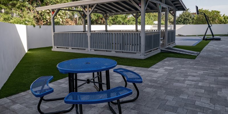 A peaceful outdoor area with a shaded gazebo, blue picnic table, and basketball court, offering a perfect spot for relaxation and activities.