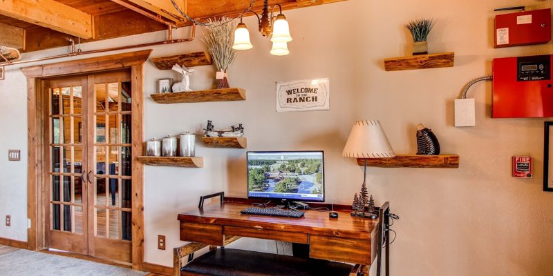  A rustic, cozy office nook featuring a wooden desk with a computer, floating shelves with décor, and a "Welcome to the Ranch" sign, offering a warm workspace.