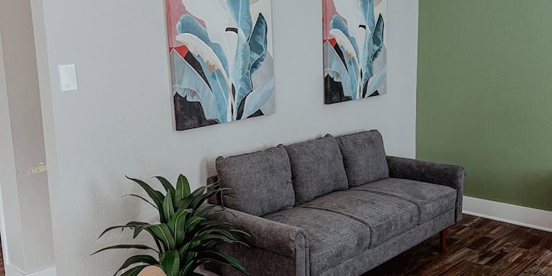 A cozy lounge area featuring a gray sofa, light tan chair, plant, and abstract artwork, creating a calm and welcoming environment with natural tones.