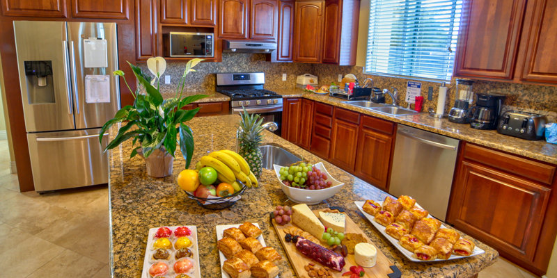 A bright, inviting kitchen featuring polished granite countertops, fresh fruits, pastries, and a beautifully arranged charcuterie board.