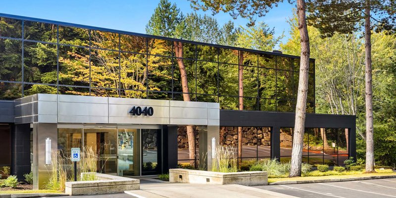  A sleek, modern building with mirrored glass reflecting lush trees, featuring a clean, welcoming entrance surrounded by nature.