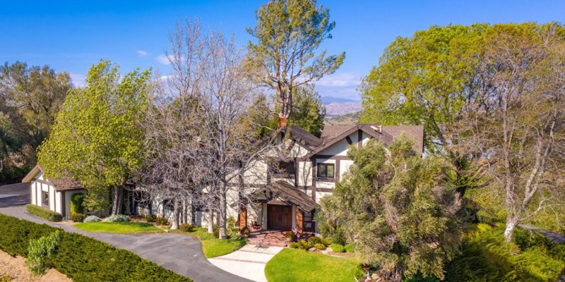 A charming, two-story home surrounded by lush trees and greenery, with a winding driveway, set against a backdrop of scenic hills and blue skies.