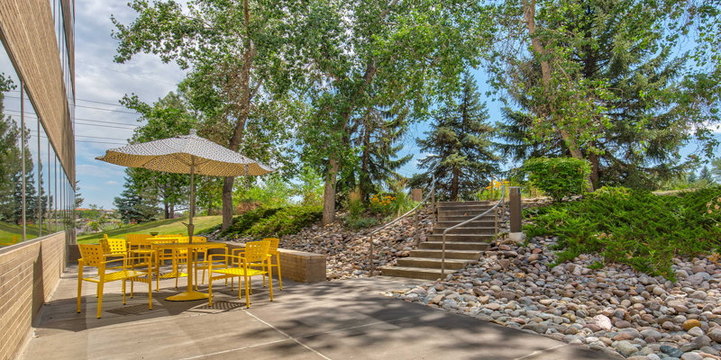  A bright outdoor seating area with yellow chairs and a matching umbrella provides a peaceful, shaded spot near greenery and stone landscaping.