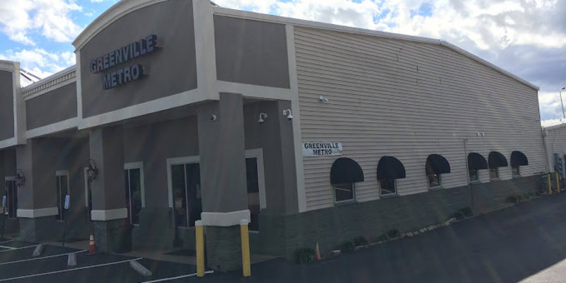 A modern building labeled "Greenville Metro" with large windows and black awnings. The parking lot is spacious, and the sky is bright and clear.