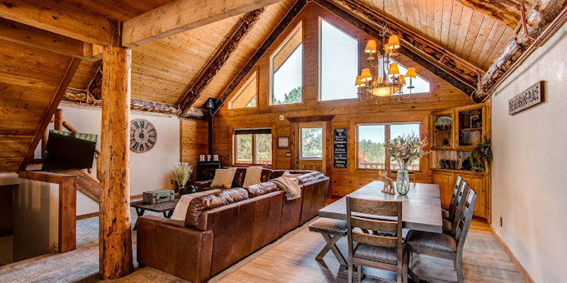 A warm, inviting cabin interior with vaulted ceilings, large windows, rustic wooden beams, a cozy living area, and a dining table, creating a charming retreat.