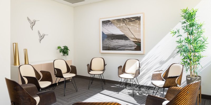 A bright and inviting therapy room with comfortable chairs arranged in a circle. Natural light streams in through large windows, complementing the calming decor, including tasteful artwork, minimalist plants, and warm, neutral tones.