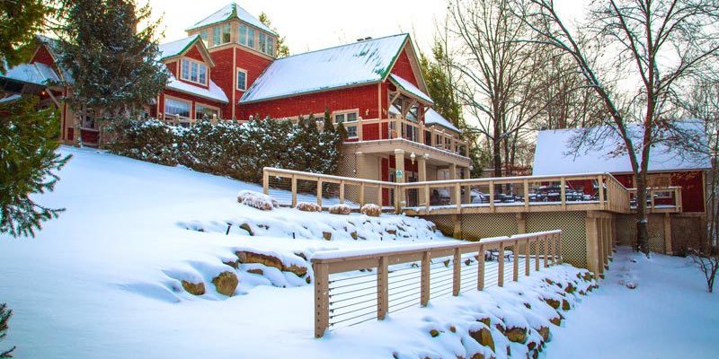 A charming red house sits amidst a snowy landscape, surrounded by trees. A wooden deck and railings lead down to the snow-covered ground.