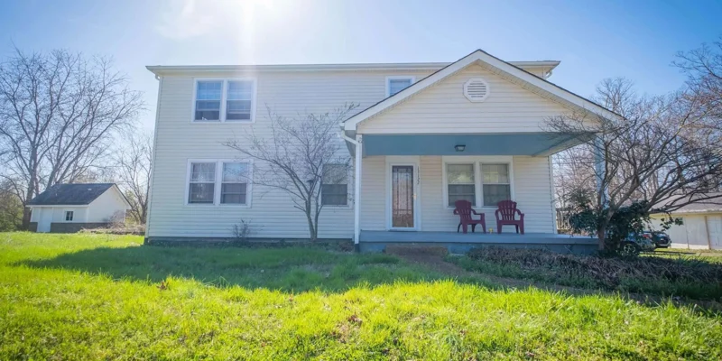  A charming two-story building with a sunny front porch, red chairs, and a lush green lawn, creating a welcoming and peaceful atmosphere.