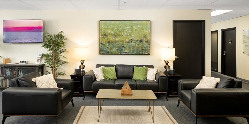 A comfortable lounge area with black leather sofas, green accent pillows, and warm lighting. Art and plants create a calm, inviting environment.