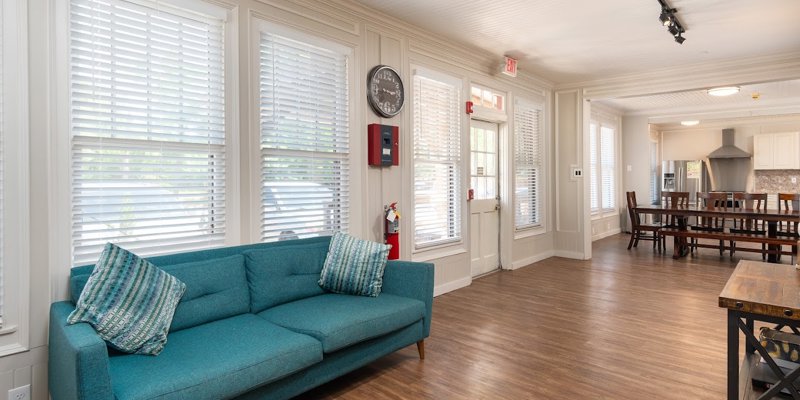 A cozy lounge area featuring a teal sofa and bright natural light from large windows, with an adjacent spacious dining and kitchen area.