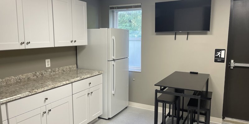  A small, clean kitchenette with white cabinets, a refrigerator, a compact table with seating, and a mounted TV, offering a cozy space for breaks.