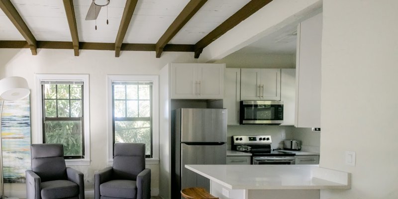 A cozy living space with two modern gray chairs, a sleek kitchen area featuring stainless steel appliances, and a rustic beamed ceiling.