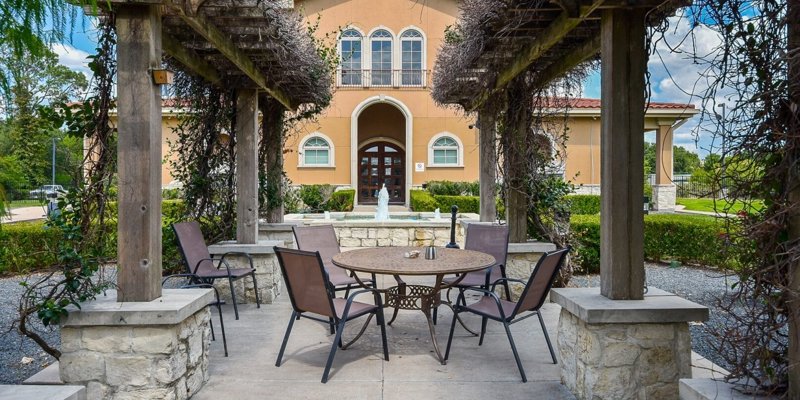 A peaceful outdoor seating area under a wooden pergola, surrounded by greenery. A charming fountain and Mediterranean-style building create a serene atmosphere.