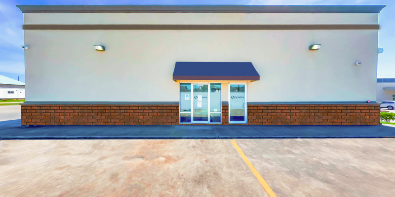 A clean, simple building entrance with glass doors under a dark awning. The exterior features light walls with a brick base and accessible parking.