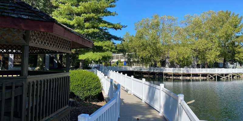 Inviting boardwalk lined with white railings, leading past a charming gazebo and serene lakeside, surrounded by lush greenery and clear skies.