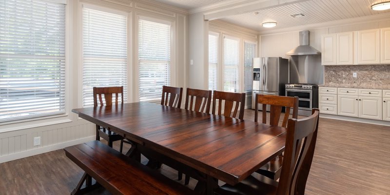  A spacious dining area with a large wooden table, bench seating, and natural light from wide windows, creating a welcoming atmosphere for gatherings.