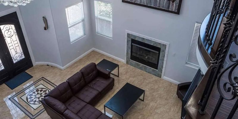 A spacious, elegant living room with high ceilings, a large sectional sofa, and a fireplace. A modern chandelier and world map artwork add sophistication to the well-lit, open space.