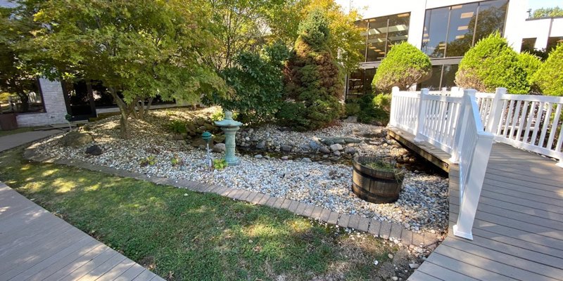 A tranquil garden area with a small stone-lined creek, surrounded by lush greenery and a charming white footbridge under clear skies.