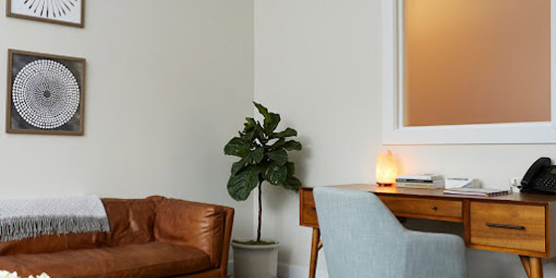 A cozy, stylish office space featuring a comfortable leather couch, modern artwork, a wooden desk, and a soft-lit lamp, creating a warm and inviting atmosphere.