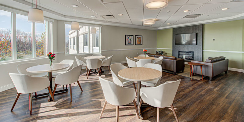 A bright, inviting dining area with stylish white chairs, round tables, large windows, and a cozy seating space with a TV and fireplace.