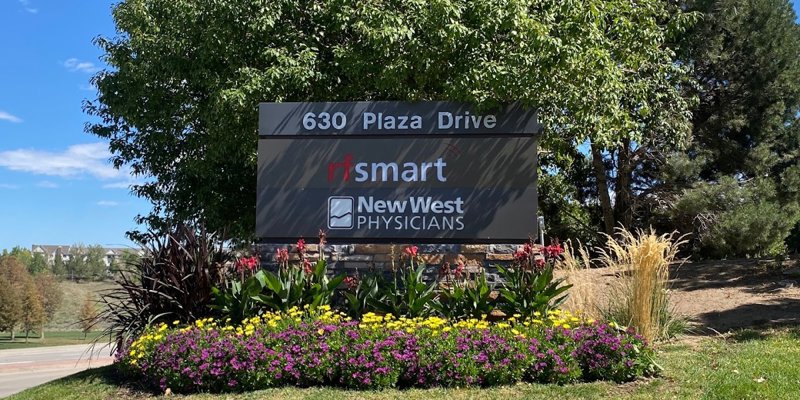 A street sign for 630 Plaza Drive, featuring businesses like rfsmart and New West Physicians, surrounded by vibrant flowers and greenery.