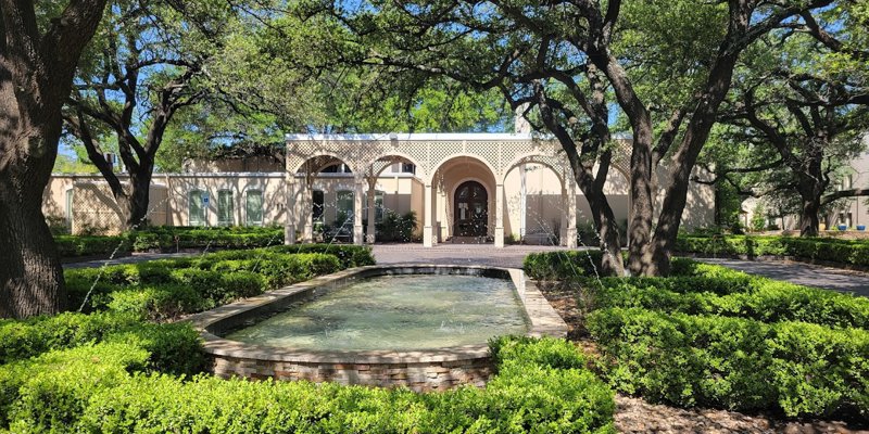 Elegant courtyard entrance surrounded by lush greenery, shaded by large oak trees, and highlighted by a fountain with arched columns.