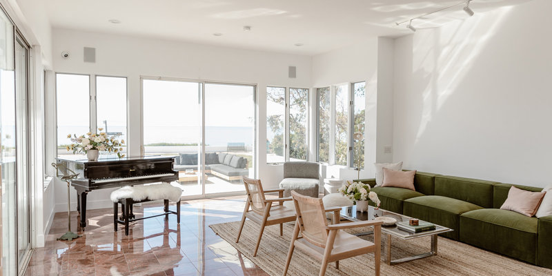 A bright, elegant living space with large windows, a piano, green sofa, modern furniture, and ocean views, creating a serene and airy atmosphere.