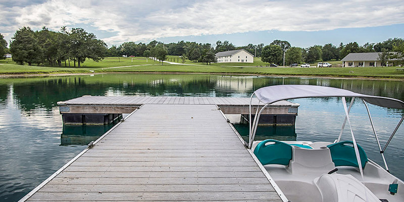 A peaceful dock with a paddleboat overlooking a serene lake, surrounded by lush green trees and distant buildings, perfect for relaxation.